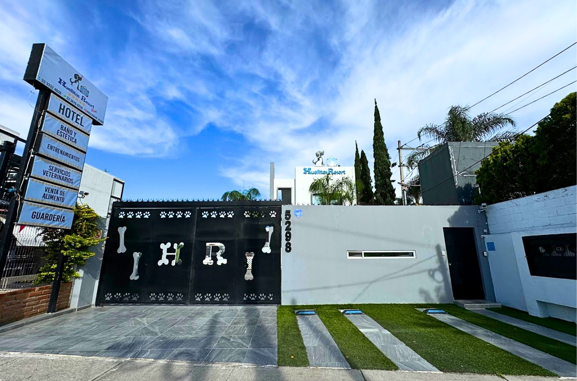 Modern black metal gate entrance with HRI signage, white contemporary building, manicured lawn with pavers, directional sign on left, blue sky